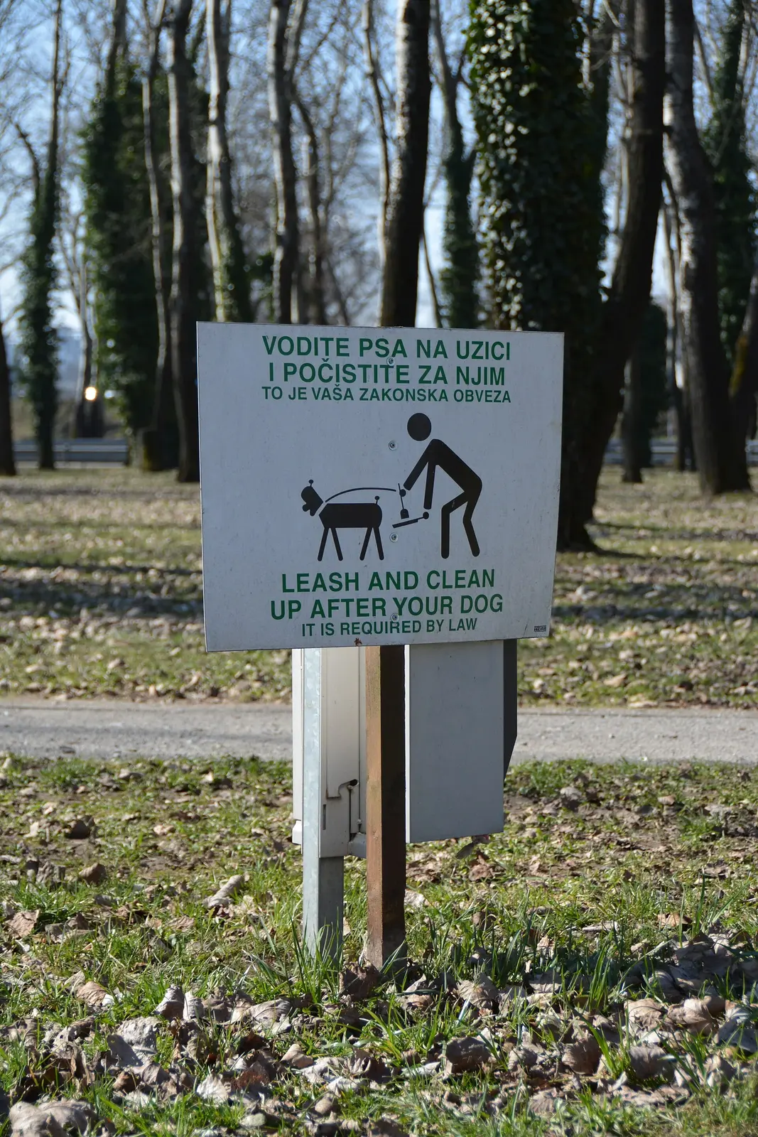 Bilingual leash and dog-waste sign beside a Bundek Park path